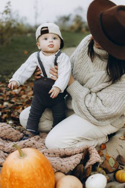 Mutlu anne ve tatlı bebek, sonbahar bahçesinde balkabağıyla rahatlıyor ve yapraklarını battaniyeye döküyor. Mutlu Şükran Günleri ve Cadılar Bayramı. Şık bir anne ve küçük tatlı bir çocuk sonbahar parkında