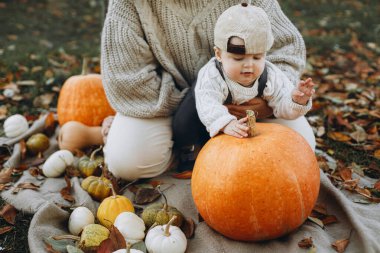 Mutlu anne ve tatlı bebek, sonbahar bahçesinde balkabağıyla rahatlıyor ve yapraklarını battaniyeye döküyor. Mutlu Şükran Günleri ve Cadılar Bayramı. Şık bir anne ve küçük tatlı bir çocuk sonbahar parkında