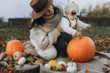 Mutlu anne ve tatlı bebek, sonbahar bahçesinde balkabağıyla rahatlıyor ve yapraklarını battaniyeye döküyor. Mutlu Şükran Günleri ve Cadılar Bayramı. Şık bir anne ve küçük tatlı bir çocuk sonbahar parkında