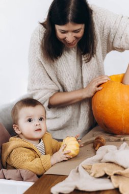 Cadılar Bayramı balkabağı oymacılığı. Mutlu anne ve tatlı bebek, masada hayaletler, örümcekler ve yarasalarla balkabağı kesiyor. Balkabağı feneri yapmak, Cadılar Bayramı hazırlığı