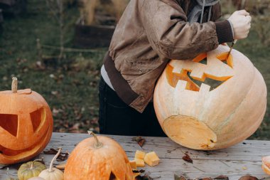Sonbahar bahçesindeki ahşap masaya cadılar bayramı balkabağı oyan adam elleri kapalı. Sonbahar hasadı. Balkabağı kesmek, balkabağı feneri yapmak. Cadılar Bayramı hazırlığı