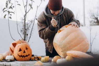Sonbahar bahçesindeki ahşap masaya cadılar bayramı balkabağı oyan şık bir adam. Sonbahar hasadı. Balkabağı kesmek, balkabağı feneri yapmak. Cadılar Bayramı hazırlığı