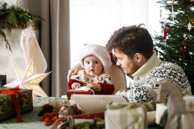Mutlu Noeller! Noel Baba ve küçük oğlu Noel Baba şapkasıyla ve şık süveterleriyle modern dekore edilmiş ağacın arka planında Noel hediyelerini birleştiriyorlar. Neşeli aile zamanı