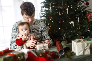 Mutlu Noeller! Mutlu baba ve bebek oğul şık süveterlerle modern dekore edilmiş ağacın arka planında noel hediyelerini birleştiriyorlar. Mutlu aile zamanları, mutlu tatiller.
