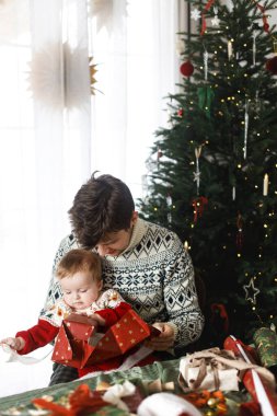 Mutlu Noeller! Mutlu baba ve bebek oğul şık süveterlerle modern dekore edilmiş ağacın arka planında noel hediyelerini birleştiriyorlar. Mutlu aile zamanları, mutlu tatiller.