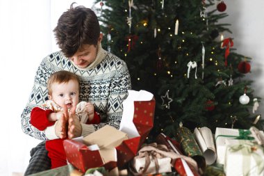 Mutlu Noeller! Mutlu baba ve bebek oğul şık süveterlerle modern dekore edilmiş ağacın arka planında noel hediyelerini birleştiriyorlar. Mutlu aile zamanları, mutlu tatiller.