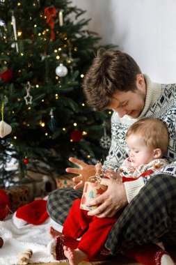 Mutlu Noeller! Mutlu bir baba ve güzel bir erkek çocuk ellerinde modern hediye kutusuyla Noel ağacının arka planında ışıklar tutuyorlar. Neşeli aile zamanları, kış tatili hazırlıkları