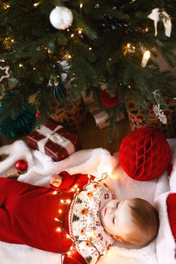 Şenlikli süveter giyen, altın ışıklarla örtülü, Noel ağacının altında hediyeli, güzel bir erkek bebek. Mutlu Noeller! Atmosferik tatil zamanı, aile süsleme ağacı.