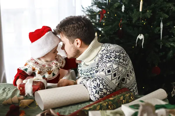 Mutlu Noeller! Noel Baba ve küçük oğlu Noel Baba şapkasıyla ve şık süveterleriyle modern dekore edilmiş ağacın arka planında Noel hediyelerini birleştiriyorlar. Neşeli aile zamanı