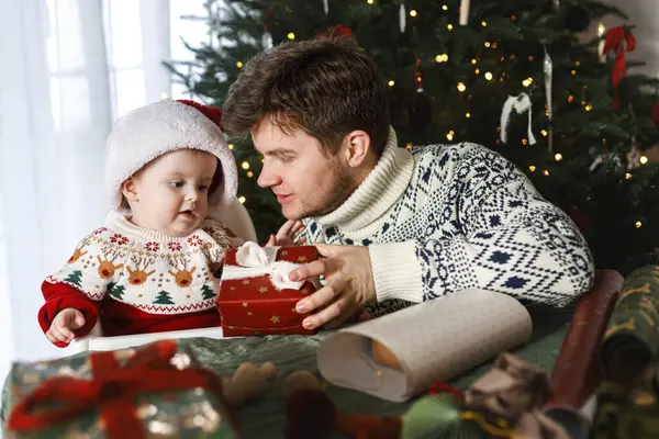 Mutlu Noeller! Noel Baba ve küçük oğlu Noel Baba şapkasıyla ve şık süveterleriyle modern dekore edilmiş ağacın arka planında Noel hediyelerini birleştiriyorlar. Neşeli aile zamanı