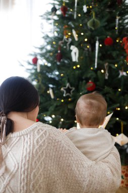 Şık bir anne şirin bir bebeğe sarılıyor ve ışıklarla süslenmiş modern İskandinav Noel ağacına bakıyor. Anne ve oğul örülmüş süveter, atmosferik tatlı an, bebek ilk Noel