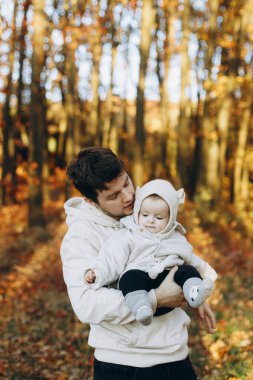 Mutlu bir baba, güz ormanında yumuşak bir akşam ışığında küçük oğluna sarılıp onu öpüyor. Babalık. Güz parkında kucaklaşan sevimli çocuklu, sıcak kazaklı şık bir adam.