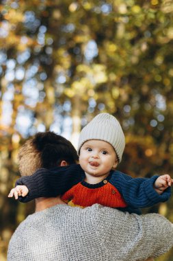 Sonbahar ormanlarında babasının ellerinde komik duyguları olan sevimli, mutlu bir bebek. Babalık. Hoş çocuklu şık bir adam rahat kazaklar içinde sonbahar parkında dinleniyor.