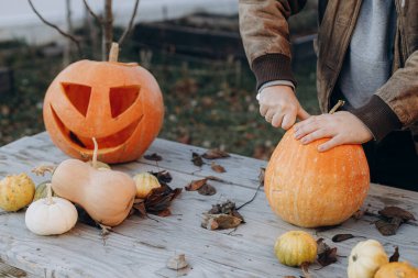 Sonbahar bahçesindeki ahşap masaya cadılar bayramı balkabağı oyan adam elleri kapalı. Sonbahar hasadı. Balkabağı kesmek, balkabağı feneri yapmak. Cadılar Bayramı hazırlığı