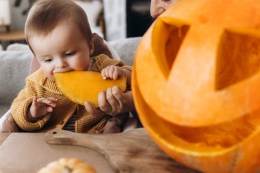 Mutlu Cadılar Bayramı. Tatlı bebek balkabağını ısırıyor ve anne Cadılar Bayramı dekoruyla masaya balkabağı feneri oyuyor. Aile Jack O feneri yapıyor, bayram zamanı