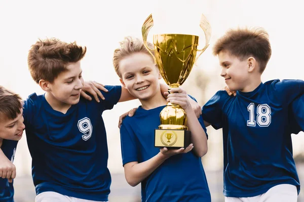Happy young boys in sports team winning a soccer-football game and holding a golden trophy. Kids soccer players celebrate a winning in school sports tournament. Happy boys in a football team