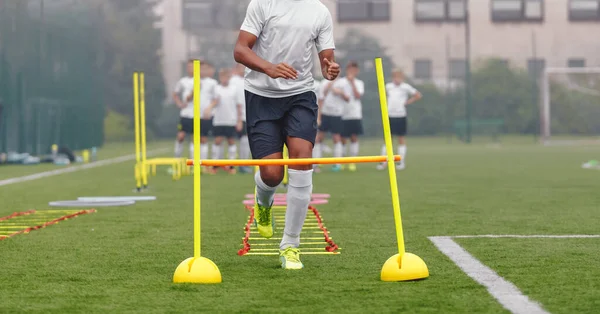 Teenage african player running at practice drill. Soccer teenager jumping over training ladder and hurdles. Football teeam in blurred background