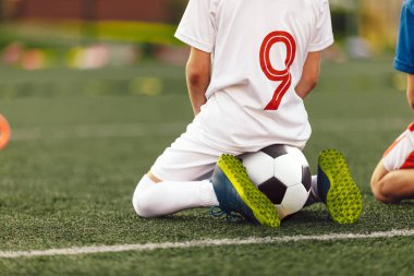 Genç beyaz futbolcu futbol sahasında oturuyor. Spor antrenmanında antrenman izleyen küçük çocuklar. Futbol formalı çocuklar.