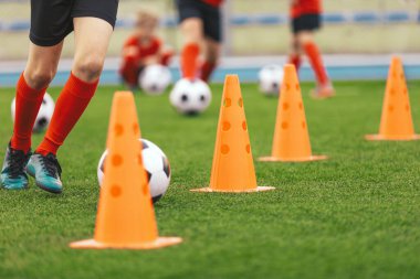 Bir sıra turuncu futbol antrenman konisi. Oyuncular antrenman konileri arasında futbol toplarıyla koşuyorlar. Çocuk oyuncular için futbol eğitimi kursu