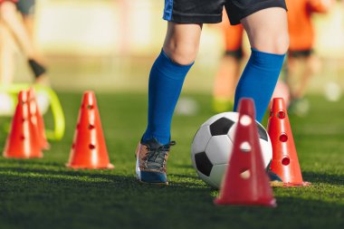 Çocuklar için futbol antrenmanı. Çocuklar slalom taliminde klasik futbol topunu tekmeliyor. Okullu çocukların bacakları hızlı koşuyor ve kırmızı antrenman konilerinin arasında futbol topunu tekmeliyor. 