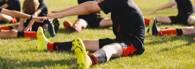 Okul çocukları çimen spor sahasında esniyorlar. Siyah futbol formalı futbol oyuncuları antrenmandan sonra bacaklarını esnetiyorlar. Spor takımı dışarıda antrenman yapıyor. 