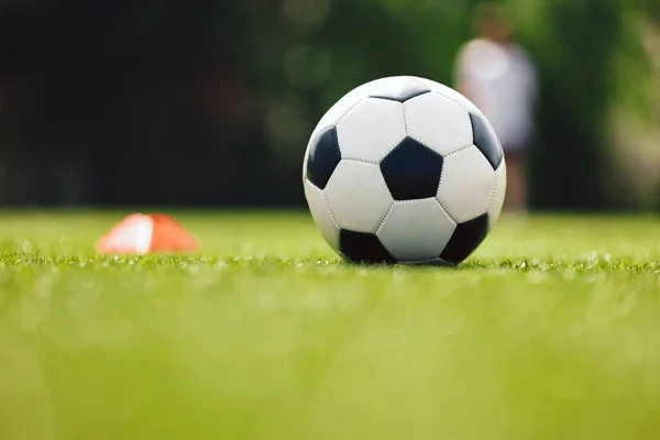Soccer Ball and Red Cone Marker on Training Pitch. Football Turf. Soccer Stadium in Blurred Background. Young Players Practicing in the Background