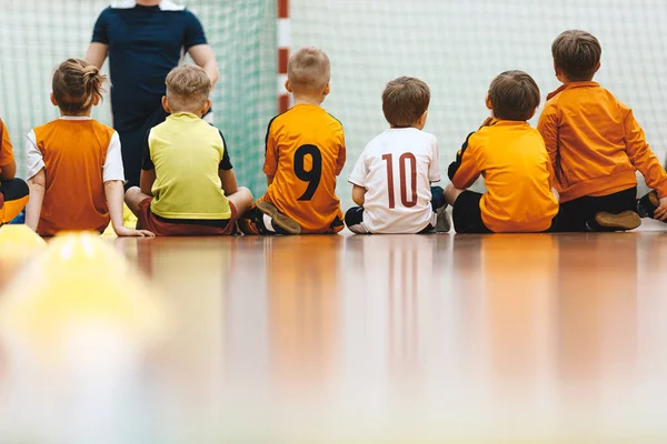 Physical education class for elementary-age children. Kids sitting on the wooden court and listening to coach trainer. School kids in sports jersey uniforms in training