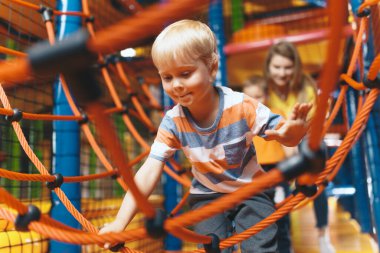 Eğlence parkında bir dadıyla oynayan mutlu çocuklar. Heyecanlı çocuklar ağ iplerinde birlikte oynuyorlar. Bir alışveriş merkezinin renkli oyun bahçesinde oynayan sevimli okul çocukları.