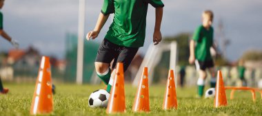 Slalom antrenmanında top sürüyor. Okul çocukları için futbol kampı. Çim sahada futbol antrenmanı ekipmanları