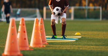 Futbolcu Çocuk Topu Ellerinde Tutuyor ve Engelli Yoldan Zıplıyor. Sports Agility Track 'i engelliyor. Çocuk Hız ve Çeviklik Yeteneklerini Geliştiriyor