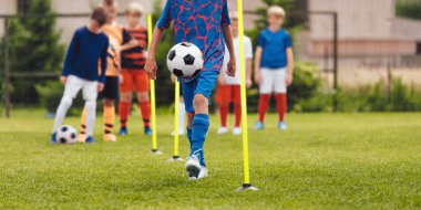 Futbol hokkabazlığı antrenmanı. Liseli çocuk antrenman sahasında direkler arasında futbol topuyla hokkabazlık yapıyor. Formalı mutlu bir çocuk spor giyiniyor ve futbol oynuyor.