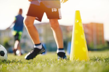 Futbol oyuncusunun bacakları antrenman konisinin yanında top sürüyor. Düşük açılı futbol antrenmanı görüntüsü. Futbol sahasında Çocuklar Egzersiz Yapıyor