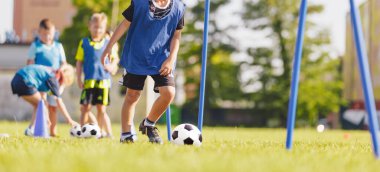 Futbol Talimleri: Slalom Matkabı. Gençlik futbolu antrenmanları. Genç oyuncular sahada antrenman yapıyor. Gençler için futbol slalom talimi. Mavi formalı çocuk külahlar arasında topla koşuyor.