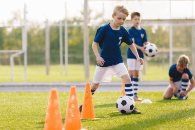 Futbol antrenmanındaki çocuklar. Okul sahasında futbol antrenmanına katılan çocuklar. Beden eğitimi sınıfındaki çocuklar. Mavi üniformalı genç oyuncular koni etrafında futbol toplarını tekmeliyorlar.
