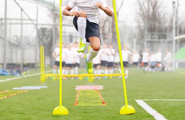 Futbol antrenmanları: oyuncu direğin üzerinden atlıyor. Gençlik futbolu antrenmanları. Genç futbolcu sahada antrenman yapıyor. Futbol merdiveni talimi. Beyaz formalı futbol takımı antrenmanda.
