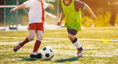İki okul çocuğu bir oyunda futbol topunu tekmeliyor. Futbolcu çocukların düellosu. Güneşli bir günde Çimenli Futbol Sahasında Futbol Maçı Oynayan Çocuklar
