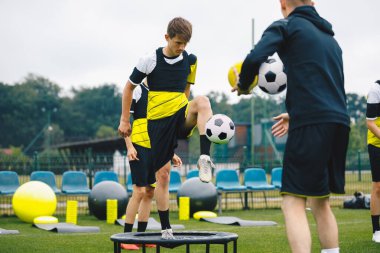 Çocuklar genç koçla futbol antrenmanı yapıyor. Futbol antrenman kampındaki gençler. Ortaokul sporcuları açık hava antrenmanında futbol becerilerini geliştiriyorlar. Trambolinde duran oyuncu koçluk yapmak için futbol topuna tekme atıyor. 