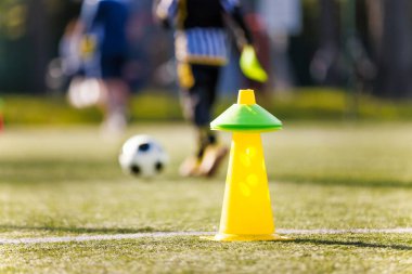 Yeşil kalemli sarı bir eğitim konisi bir futbol sahasında oturur. Arka planda, bir çocuk ve bir futbol topu dinamik eğitim oturumunda.