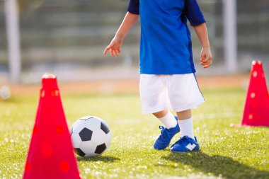 Mavi futbol kıyafeti giymiş küçük bir çocuk güneş ışığında külahta top sürüyor, koordinasyon ve ayak hareketlerine odaklanıyor.