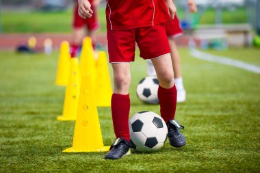 Futbol antrenmanı sırasında kırmızı futbol formalı çocuklar çimenli bir sahada sarı koni etrafında top sürme becerisi geliştiriyorlar.