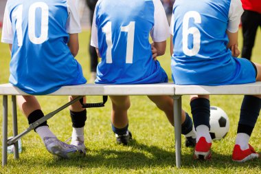 Genç Futbolcular Mavi formalarla 10, 11 ve 6 numaralarla Sunny Field 'daki Maç Molasında Bench' te oturuyorlar.