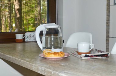 Ay çöreği ve kahve seti. Kahvaltı masasında. Arkadaki pencereden yeşil ağaçlar görünüyor.