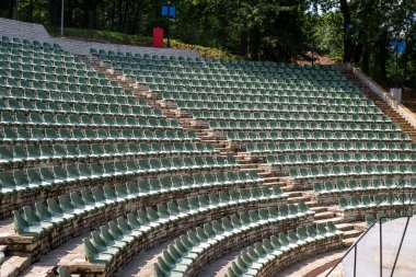 Geniş, açık hava taşlı amfi tiyatro yarı dairesel bir düzende dizilmiş sayısız yeşil plastik koltuklarla doludur.