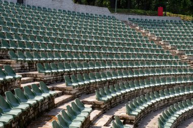 Geniş, açık hava taşlı amfi tiyatro yarı dairesel bir düzende dizilmiş sayısız yeşil plastik koltuklarla doludur.