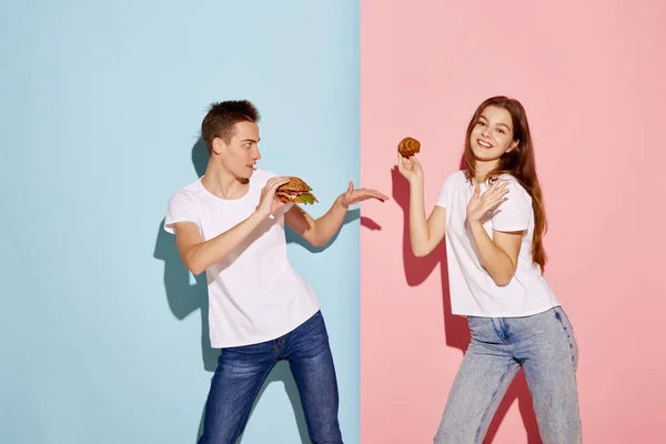 年轻人的画像 拿着汉堡包的男人和拿着羊角面包的女人在蓝色粉红的背景下摆出孤立的姿势 食物的变化 人际关系 情感的概念 广告的复制空间 — 图库照片