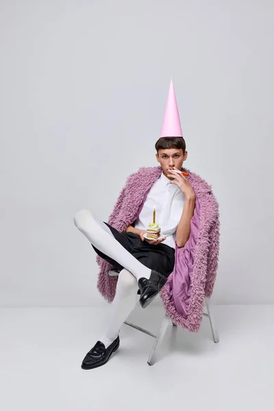 Portrait of stylish boy posing in pink furry coat with birthday cone and cake isolated over grey background. Stylish party. Concept of modern fashion, art photography, style, queer, uniqueness, ad