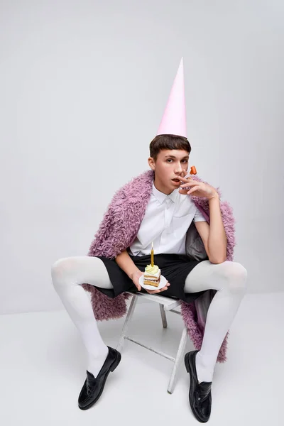 ピンクの毛皮のコートに誕生日コーンとグレーの背景に隔離されたケーキを身に着けているスタイリッシュな少年の肖像画 臨時パーティーだ 現代ファッション アート写真 スタイル クィア ユニークさ 広告のコンセプト — ストック写真