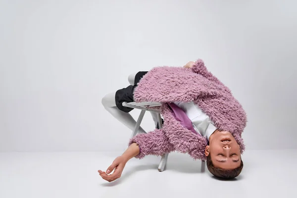 ピンクの毛皮のコートを着たスタイリッシュな少年の肖像画 灰色の背景に隔離された目で椅子に横たわっている 眠そうな気分だ 現代ファッション アート写真 スタイル クィア ユニークさ 広告のコンセプト — ストック写真