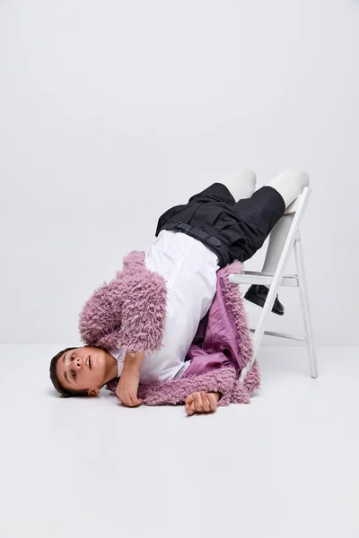ピンクの毛皮のコートにスタイリッシュな少年の肖像画 グレーの背景に隔離された椅子に横たわっている白のタイツと黒のショートパンツ 休みなさい 現代ファッション アート写真 スタイル クィア ユニークさ 広告のコンセプト — ストック写真