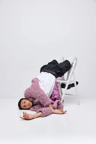 Portrait of stylish boy in pink furry coat lying on chair with legs up, posing isolated over grey background. Concept of modern fashion, art photography, style, queer, uniqueness, ad